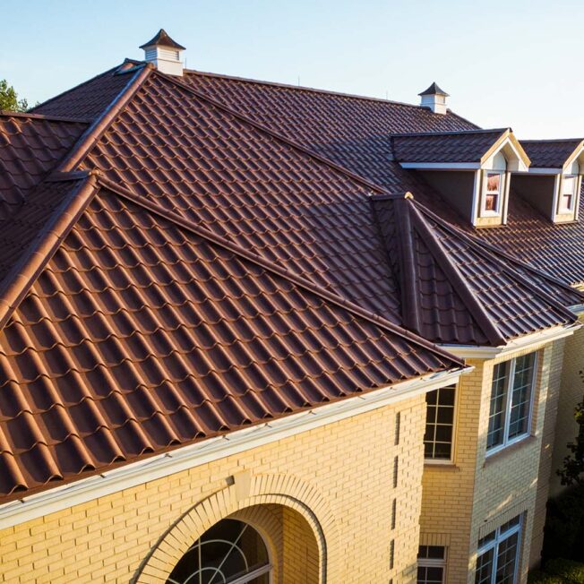 {Yellow-toned brick two-story home covered with ProVia's new metal barrel tile roof in Terracotta, example of metal roofing that looks like clay tile}