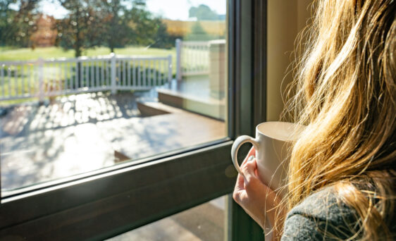{Woman holding a cup of coffee gazing out ProVia home replacement windows into her backyard, one of many vinyl replacement window types offered by ProVia}