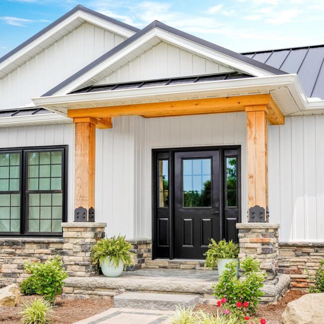 A white house with a porch with stone columns and timber and a Legacy™ Smooth 430 entry door in Coal Black with sidelites