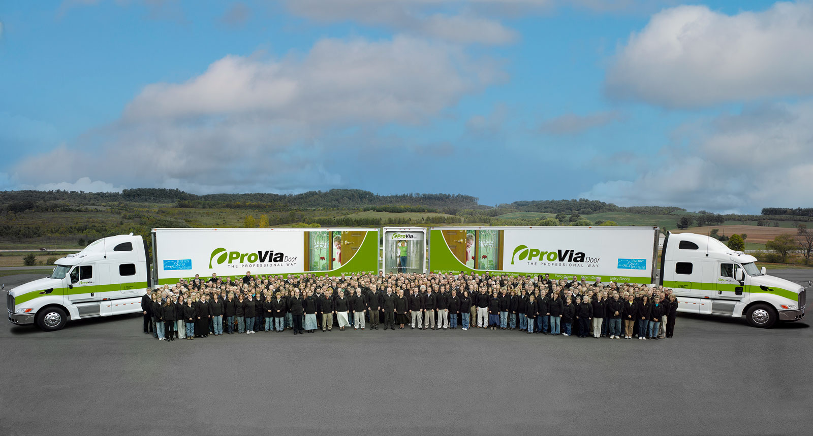 Picture of all ProVia® employees in 2007 standing in front of two ProVia semi trucks. In 2007, Precision Entry, Sugarcreek Industries, and Monarch IG were united under the name ProVia