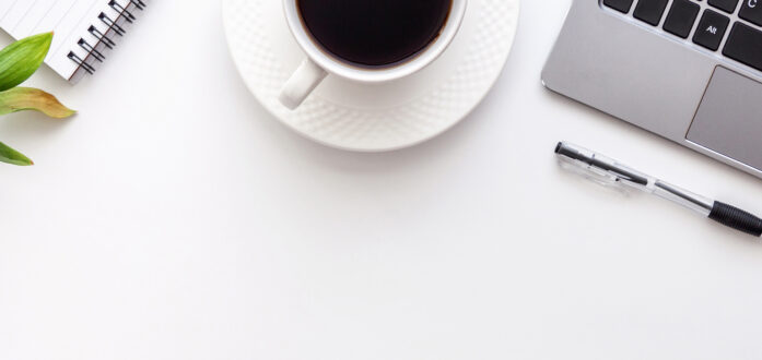 {A cup of coffee on a white saucer next to a laptop, pen, notebook, and glasses with a hint of greenery of the far left corner}