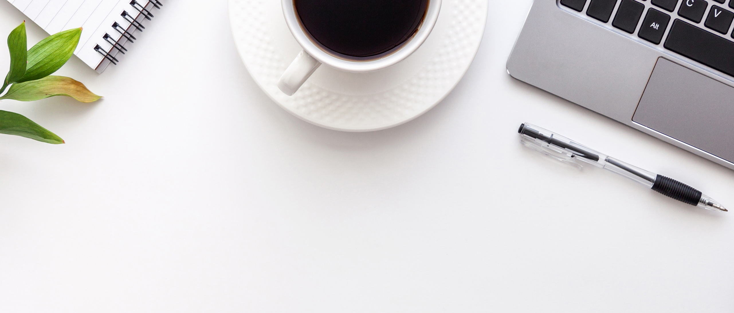A cup of coffee on a white saucer next to a laptop, pen, notebook, and glasses with a hint of greenery of the far left corner