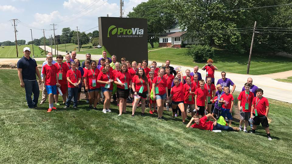 {Group of students in red shirts outside ProVia in front of the ProVia sign. These students participated in the 2017 Manufacturing Camp at ProVia.}