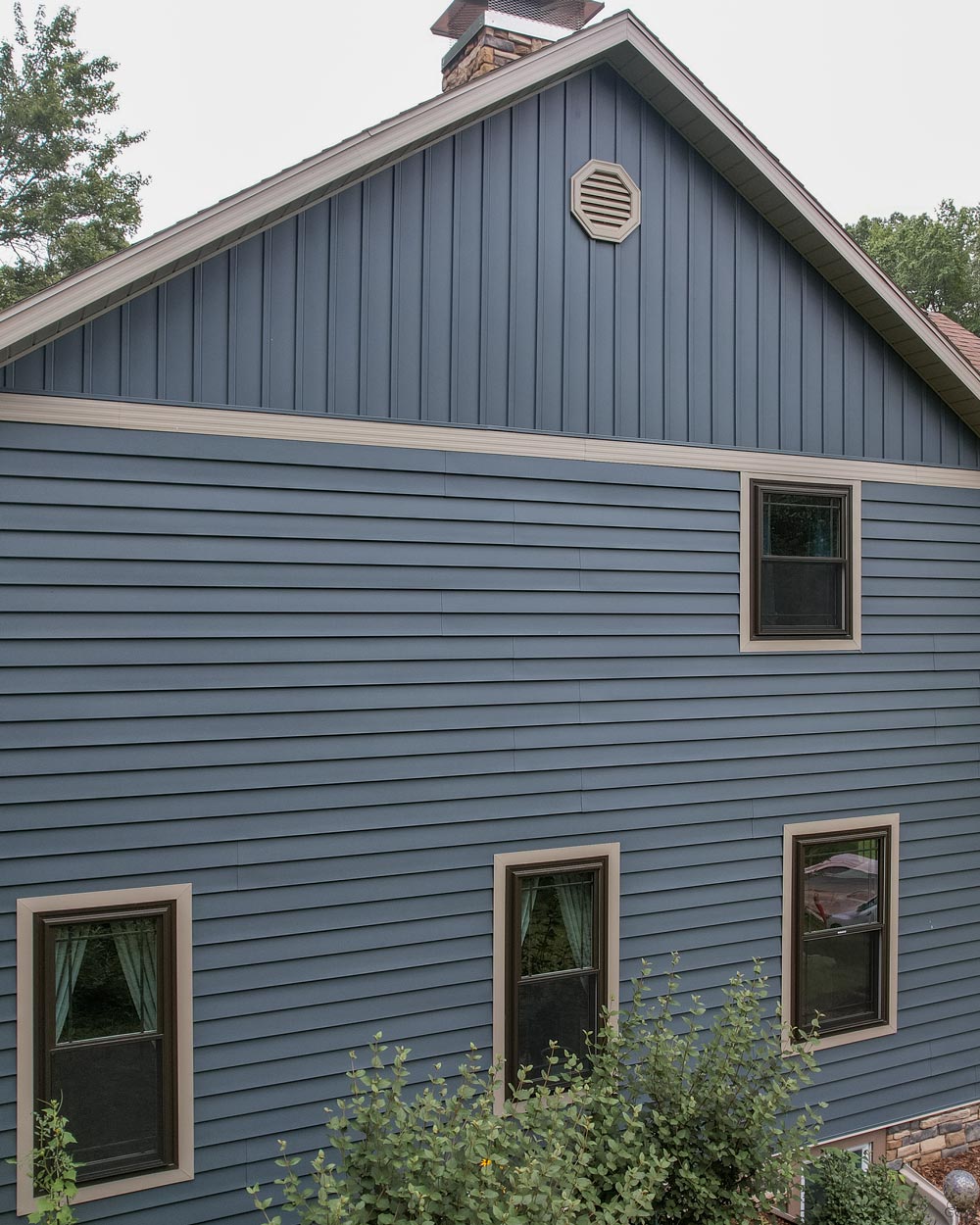Exterior view of the front of a home after getting new ProVia® CedarMAX® Insulated Vinyl Siding and Board and Batten in the color Sea Slate,  Endure™ Double Hung Windows in Classic Bronze, and Summit Terra Cut™ manufactured stone